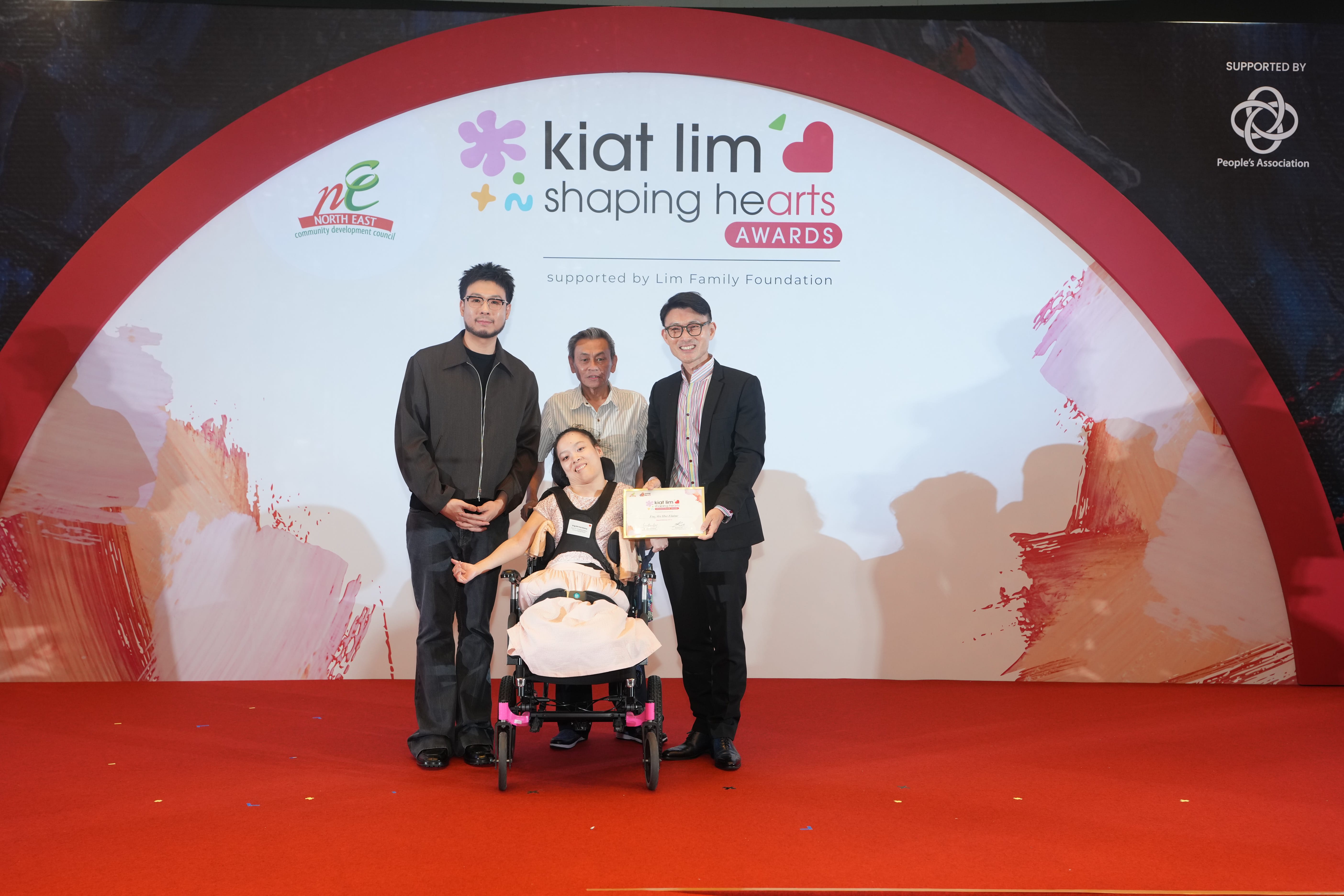 Award recipient standing on stage with the presenter, holding their certificate and posing for a group photo during the award ceremony, with a backdrop reading ‘Kiat Lim-Shaping Hearts Awards'.